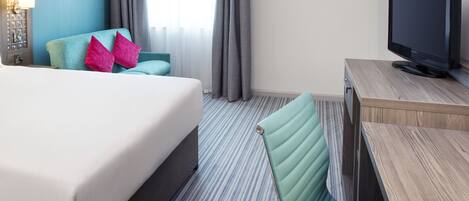 A hotel room with a bed, a desk with a television, a chair, and a window with curtains.