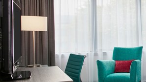 A hotel room with a wooden desk, a teal armchair with a red pillow, a standing lamp, and a large window with sheer curtains. - Leonardo Hotel Plymouth (Plymouth)