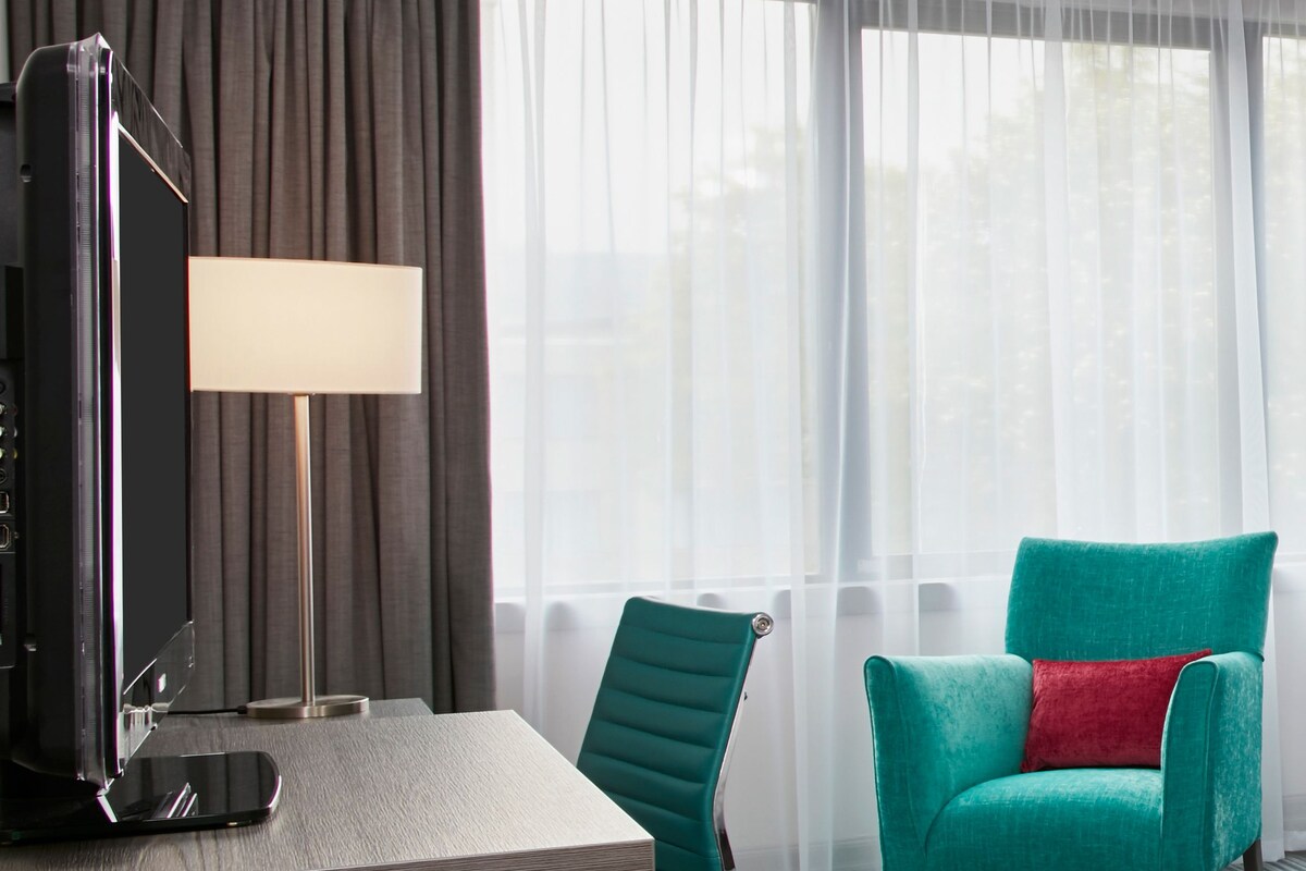 A hotel room with a wooden desk, a teal armchair with a red pillow, a standing lamp, and a large window with sheer curtains.