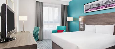 A hotel room with a large bed, a desk with a flat-screen TV, a teal chair, a cityscape painting on the wall, and a window with sheer curtains.