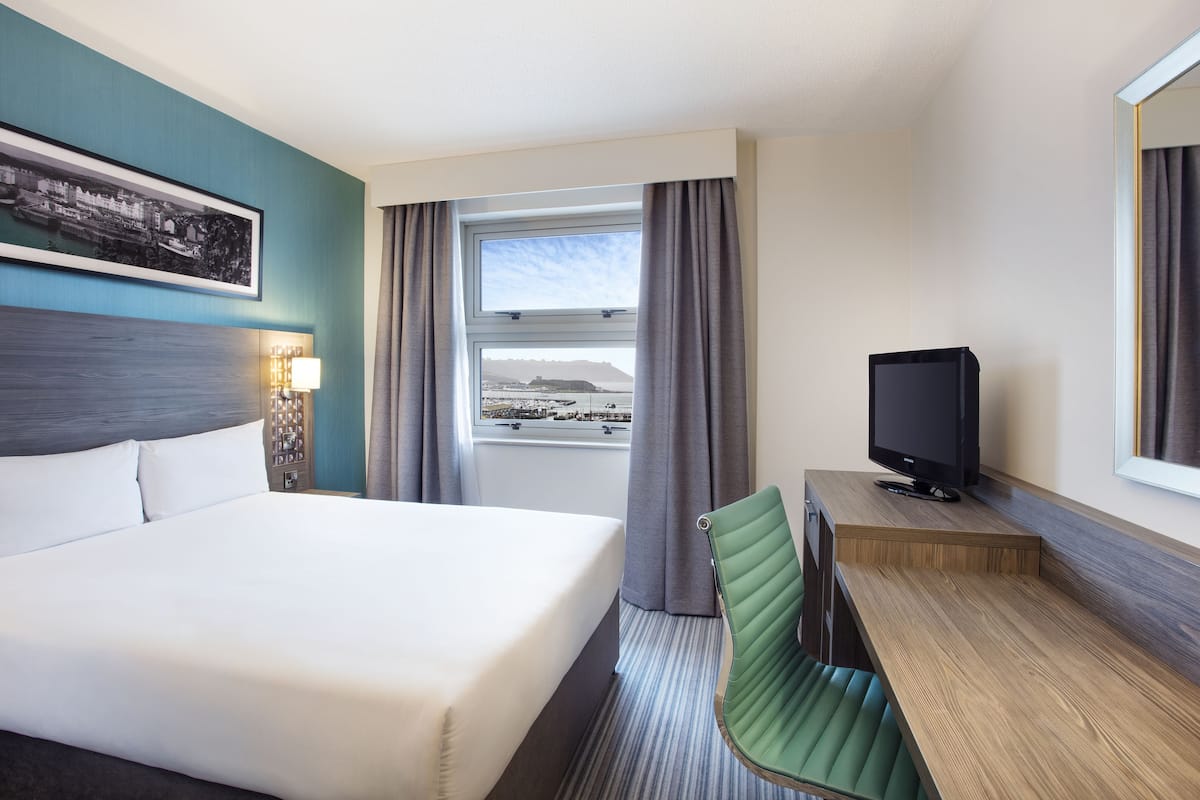 A hotel room with a bed, a desk with a television, a window with curtains, and a framed picture on the wall.