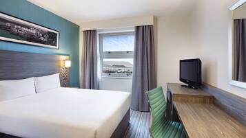 Habitación de hotel con cama, escritorio con televisor, ventana con cortinas e imagen enmarcada en la pared.