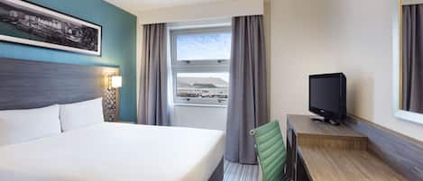 Habitación de hotel con cama, escritorio con televisor, ventana con cortinas e imagen enmarcada en la pared.
