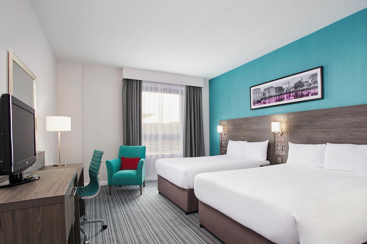 A hotel room with two beds, a desk with a TV, a teal chair, and a cityscape picture on the wall.