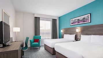 Habitación de hotel con dos camas, un escritorio con televisor, una silla color verde azulado y una fotografía de la ciudad en la pared.