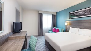 A hotel room with a bed, a desk with a TV, a chair, a window with curtains, and a wall-mounted lamp. - Leonardo Hotel Plymouth (Plymouth)