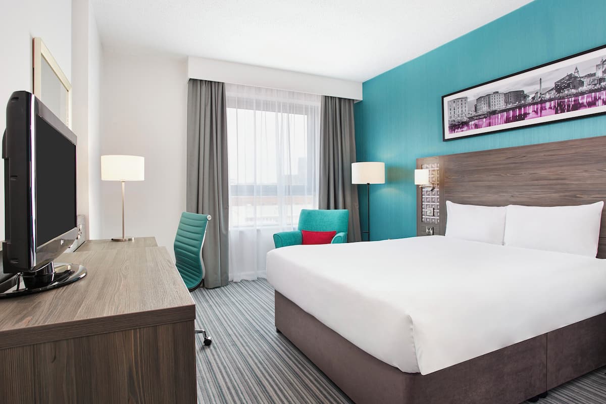 A hotel room with a large bed, a desk with a flat-screen TV, a teal chair, a cityscape painting on the wall, and a window with sheer curtains.
