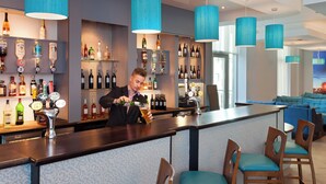 A bar area with a bartender, shelves of liquor bottles, and blue upholstered chairs. - Leonardo Hotel Plymouth (Plymouth)