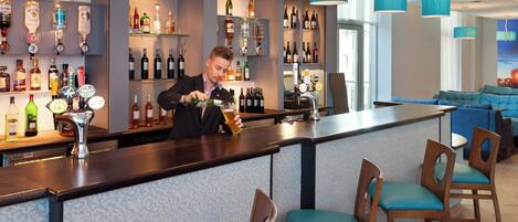 Zona de bar con un barman, estantes llenos de botellas de licores y sillas tapizadas en azul.