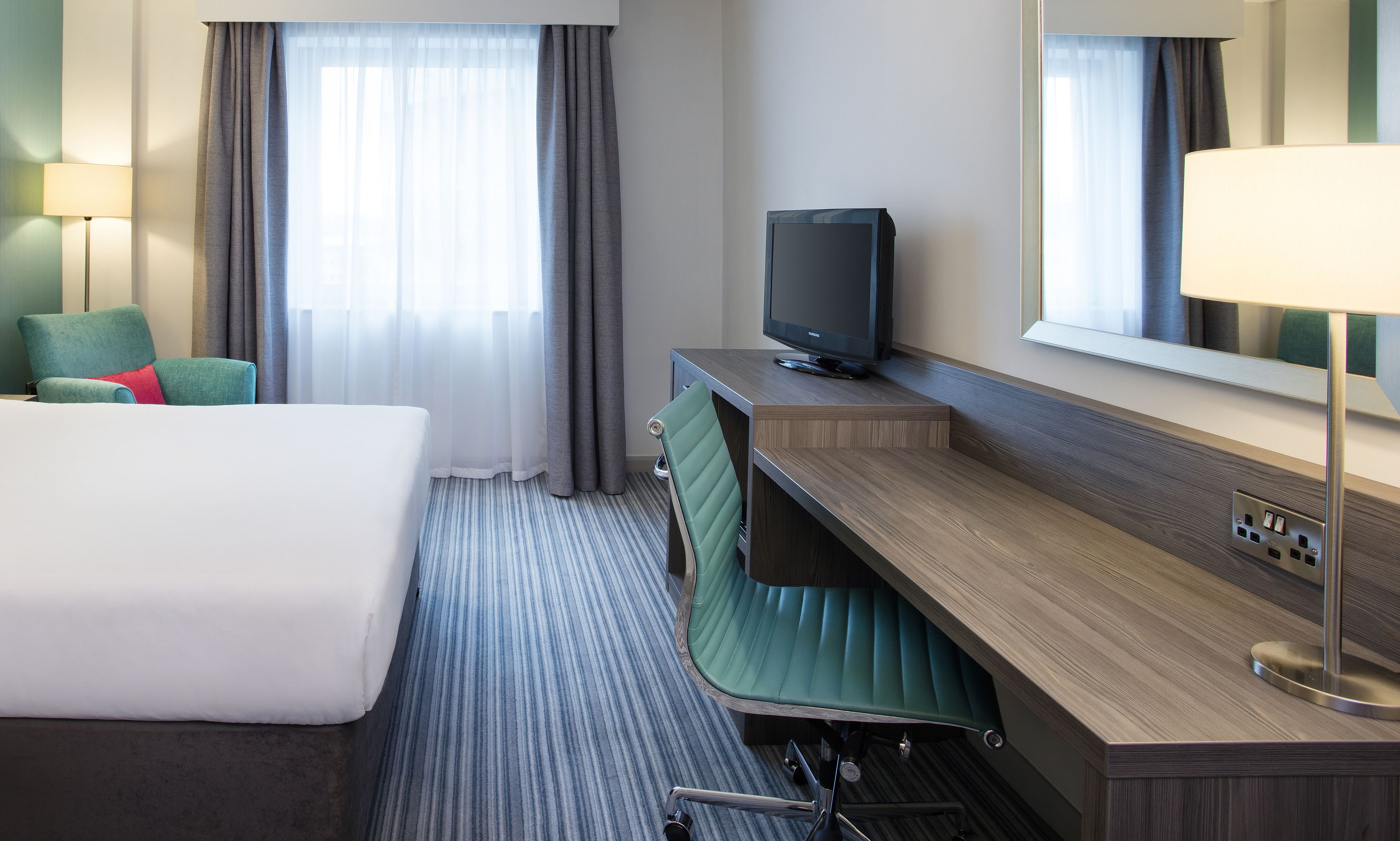 Habitación de hotel con cama, escritorio con televisor, una silla y un espejo grande.