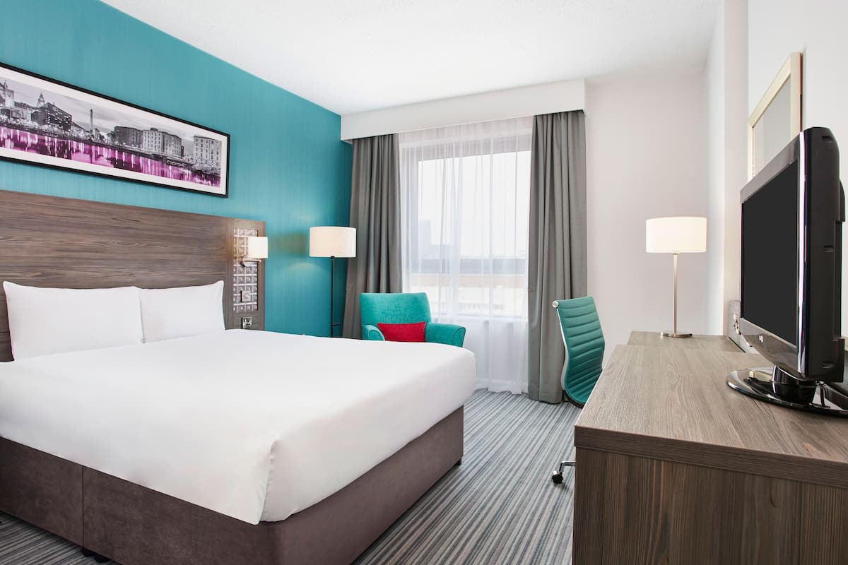 A hotel room with a large bed, a desk with a flat-screen TV, a chair, a desk lamp, and a window with curtains.