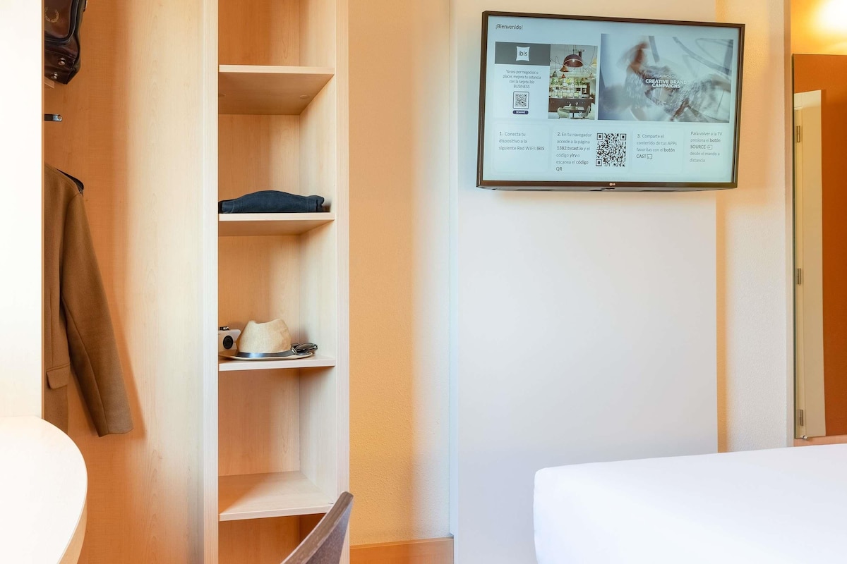 A hotel room with a bed, a desk, a chair, a wardrobe, and a wall-mounted TV displaying a welcome message.