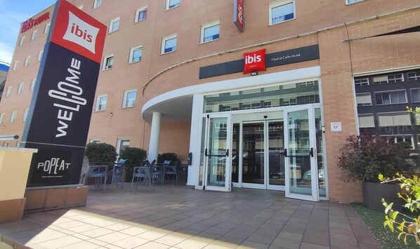 Eingang eines Ibis Hotels mit einem 'WELCOME'-Schild, Glastüren und einem Außenbereich mit Sitzgelegenheiten.
