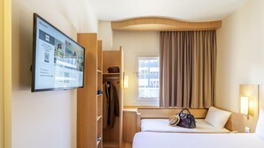 A hotel room with a bed, a desk, a wardrobe, a window with curtains, and a wall-mounted screen displaying information. - ibis Madrid Calle Alcala (Madrid)