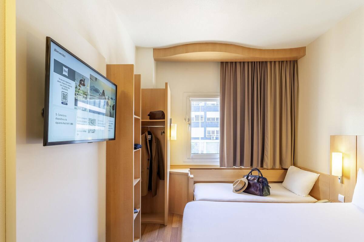 Hotelkamer met een bed, bureau, kledingkast, raam met gordijnen en een wandgemonteerde scherm met informatie.