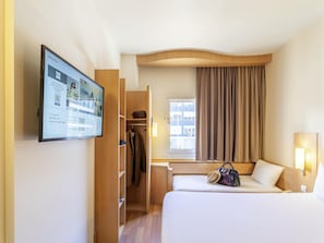Hotellihuone, jossa on sänky, työpöytä, vaatekaappi, ikkuna verhoilla ja seinälle asennettu näyttö, jossa esitetään tietoa.