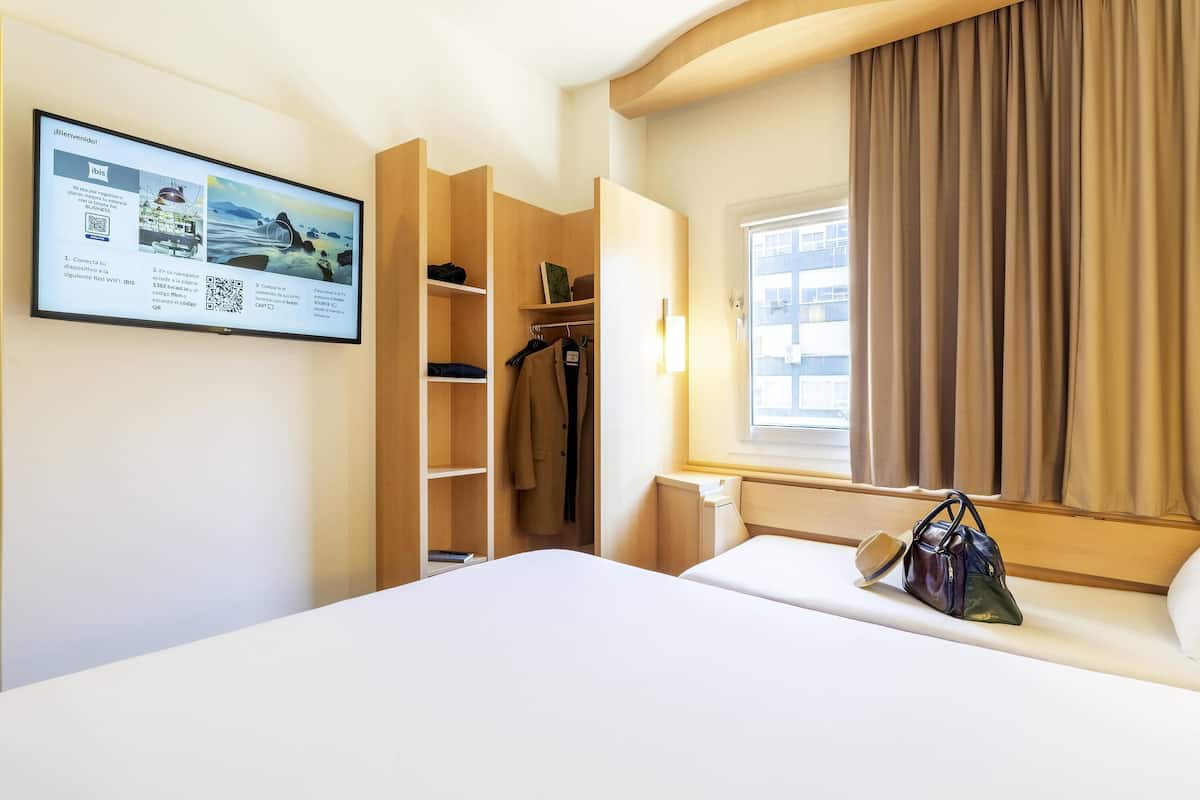 Habitación de hotel con cama, armario, ventana con cortinas y una pantalla mural mostrando un mensaje de bienvenida.