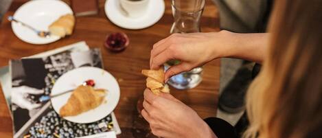 A person eating a croissant and holding a glass of water at a wooden table with a magazine.
