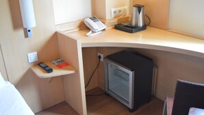 A compact hotel room with a kitchenette area, including a coffee maker, a mini-fridge, a phone, and a remote control on a small shelf.