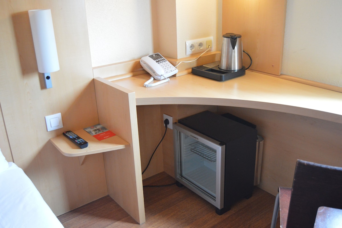 Une chambre d’hôtel compacte dotée d’un coin cuisine, comprenant une machine à café, un mini-réfrigérateur, un téléphone et une télécommande posée sur une petite étagère.