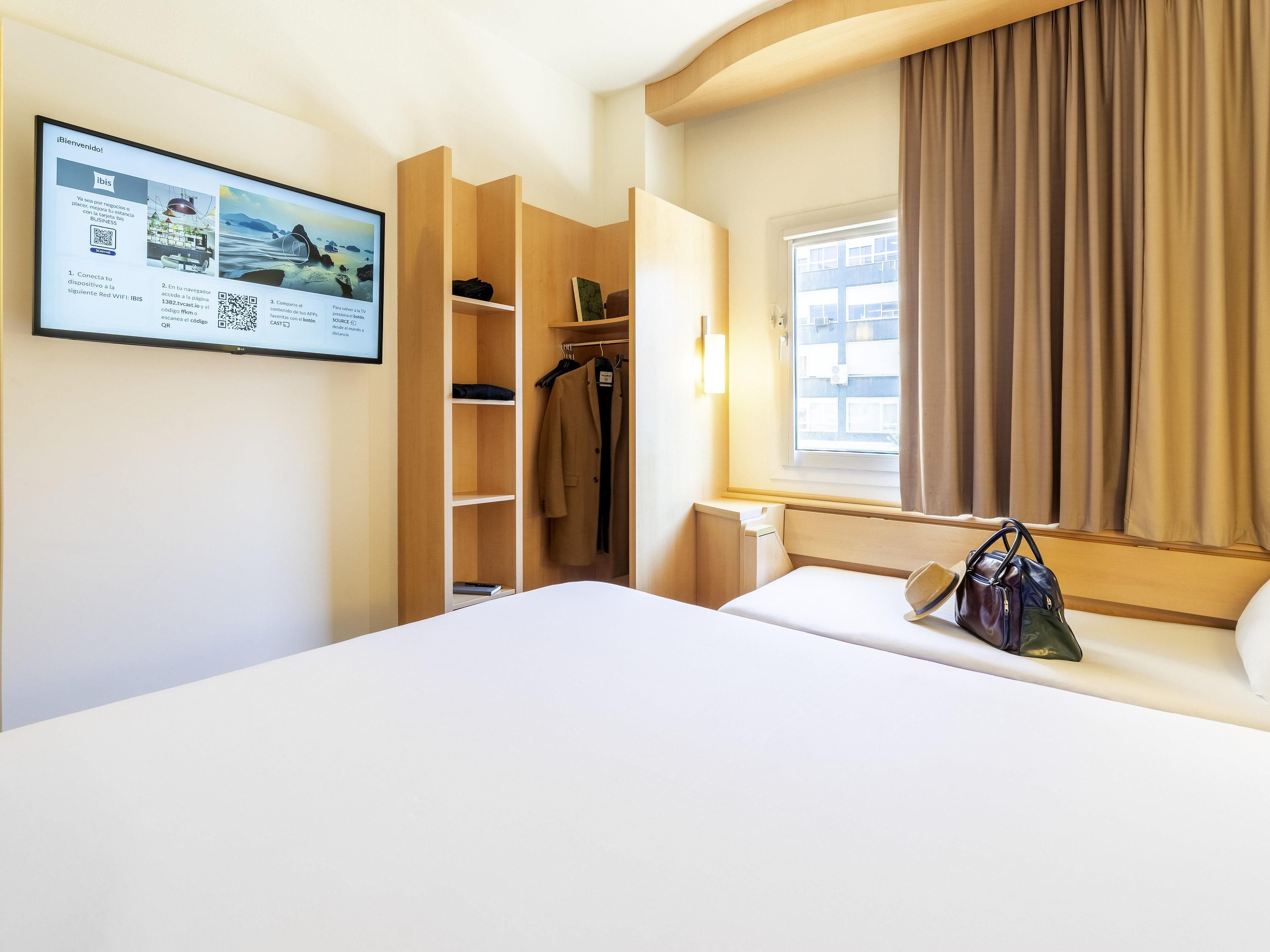 A hotel room with a bed, a wardrobe, a window with curtains, and a wall-mounted screen displaying a welcome message.