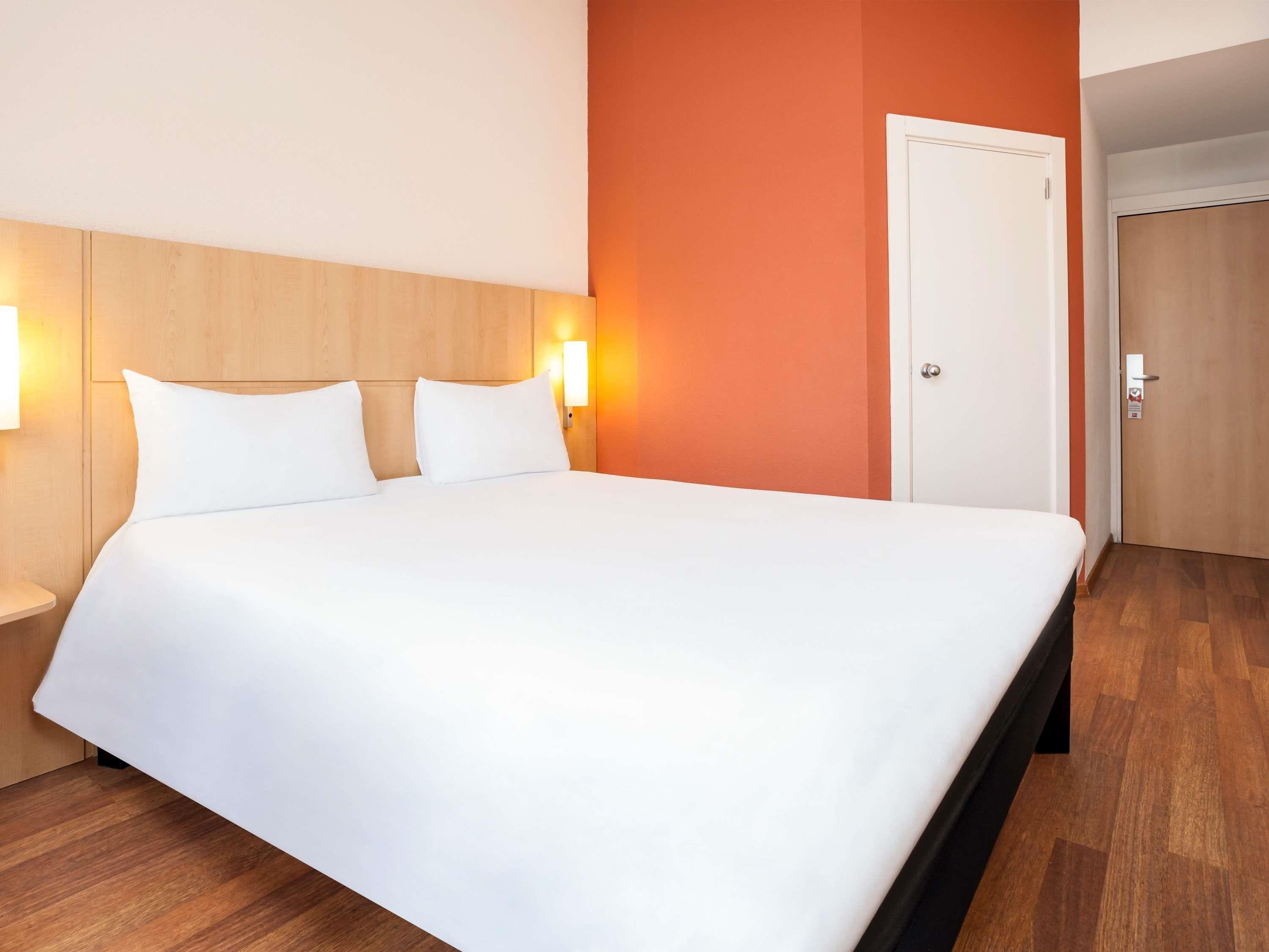 Hotellihuone, jossa on suuri sänky, puinen pääty, kaksi yöpöytävalaisinta, ovi ja puulattia.