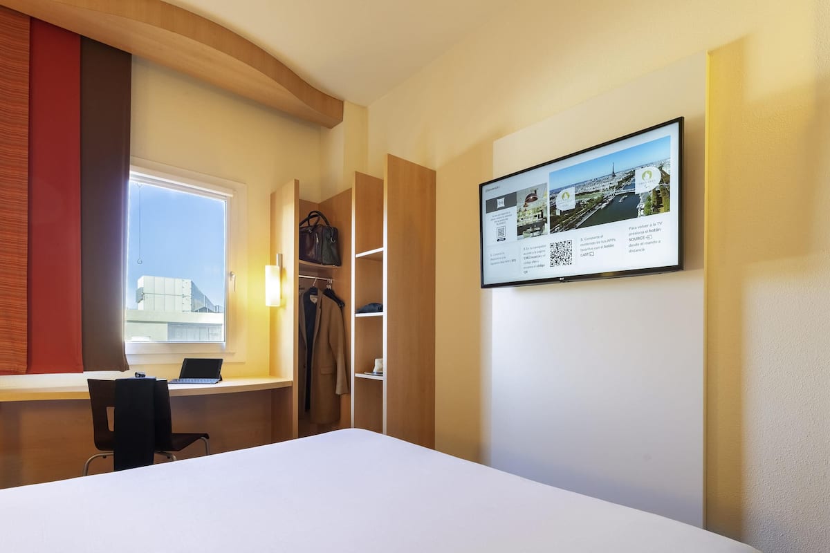 Ett hotellrum med en säng, ett skrivbord med en bärbar dator, en garderob och en väggmonterad tv som visar en stadsbild.