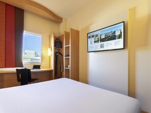 Habitación de hotel con una cama, un escritorio con una computadora portátil, un armario y un televisor de pared que muestra una vista urbana.