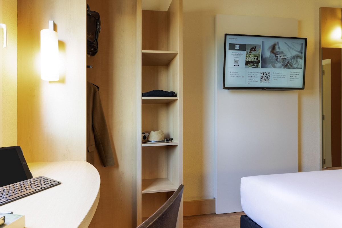 A hotel room with a desk, laptop, chair, bed, wardrobe, and a wall-mounted TV displaying a welcome message.