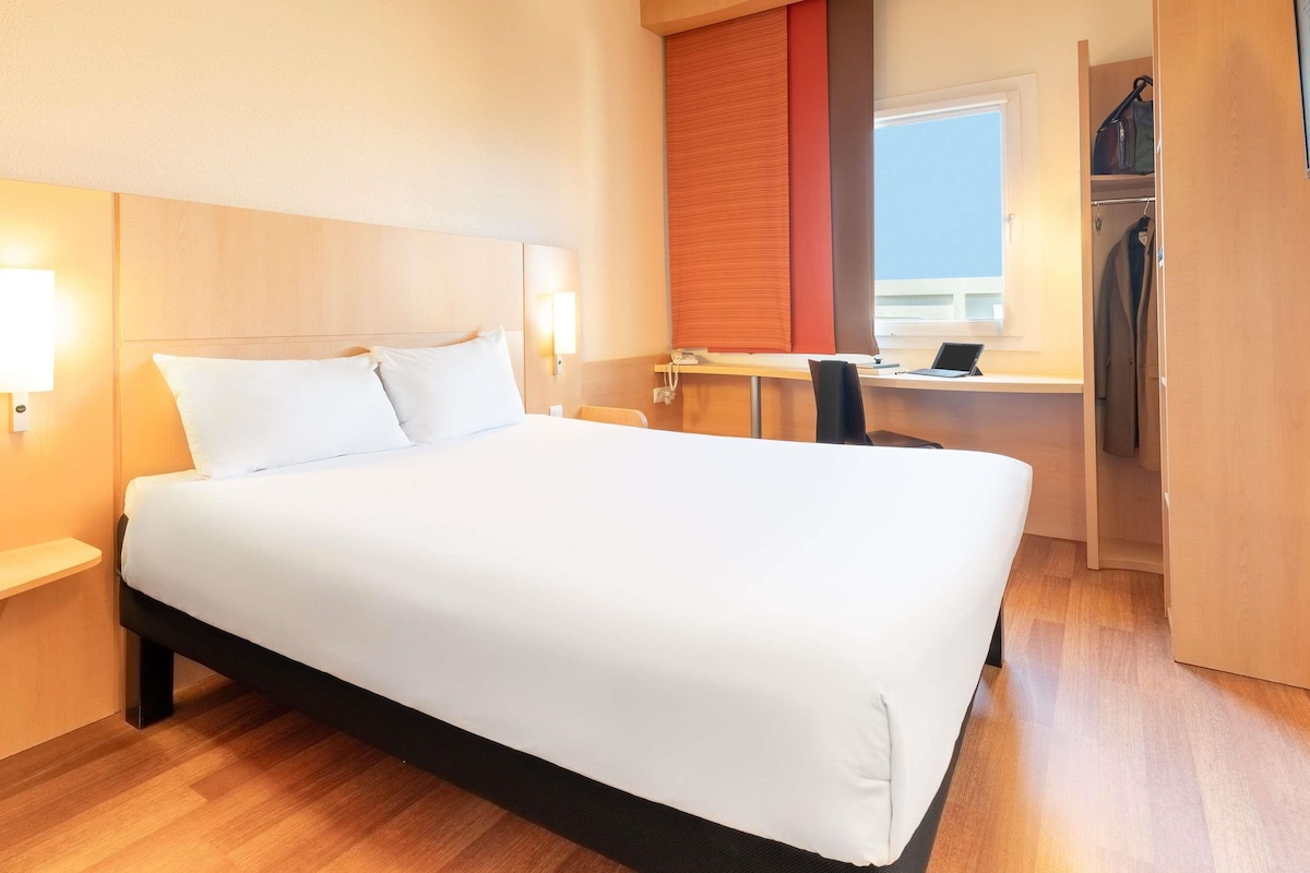 Ein Hotelzimmer mit einem großen Bett, einem Schreibtisch mit Computer, einem Stuhl und einem Kleiderschrank.