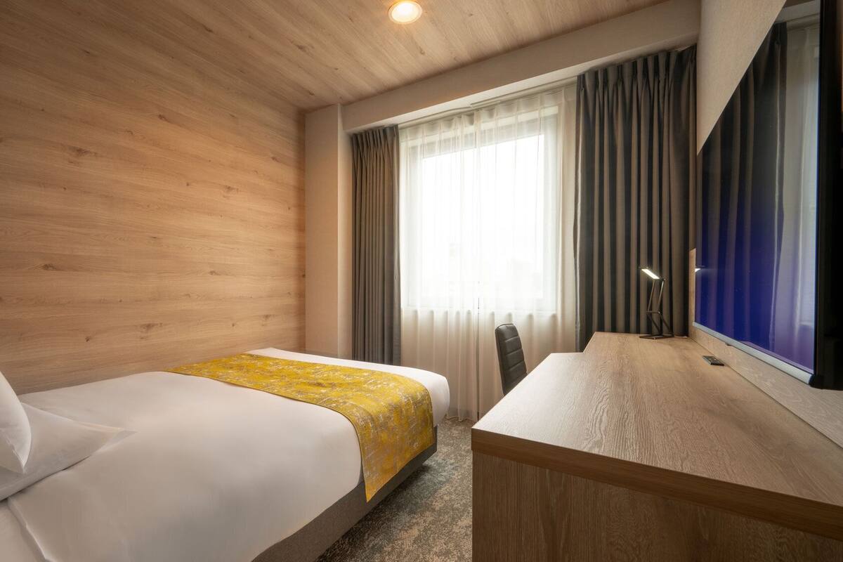 Une chambre d’hôtel avec un mur en bois, un lit recouvert d’une courtepointe blanche et jaune, un bureau avec une chaise et une grande fenêtre avec des rideaux.