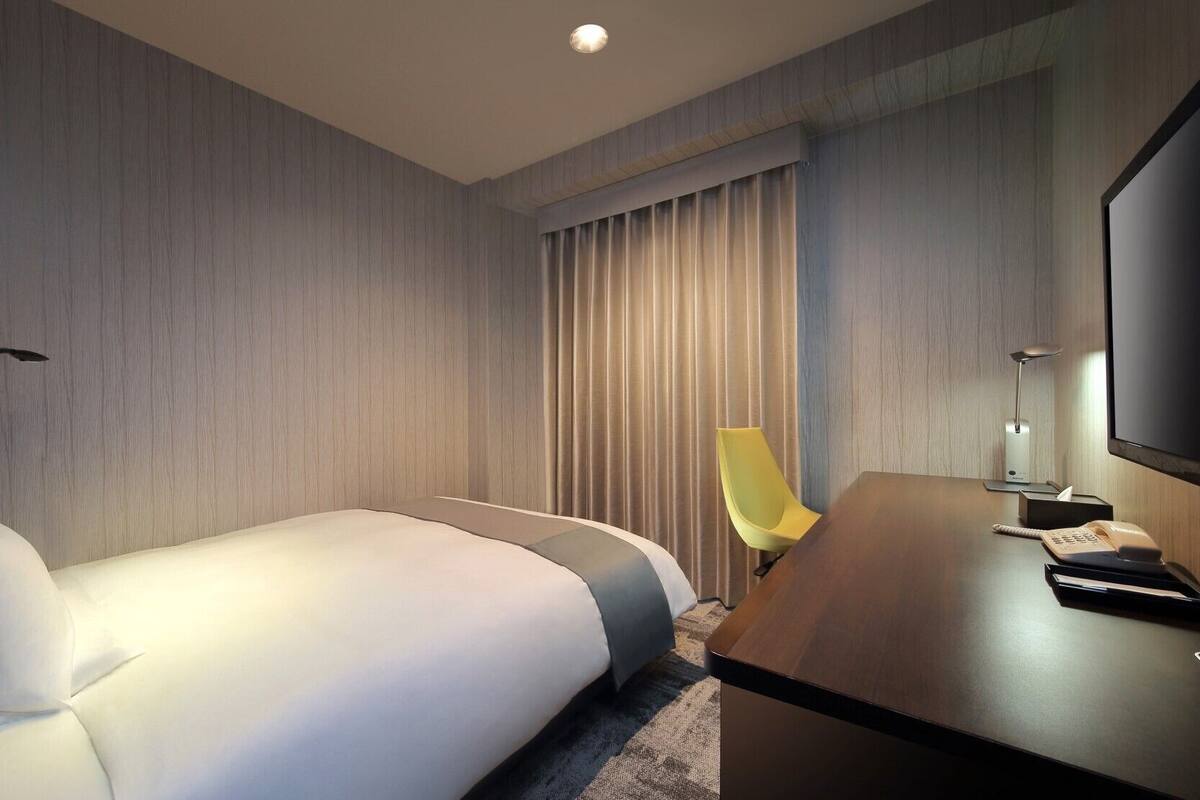 Une chambre d’hôtel moderne équipée d’un lit, d’un bureau, d’une chaise, d’une télévision et d’une fenêtre avec des rideaux.