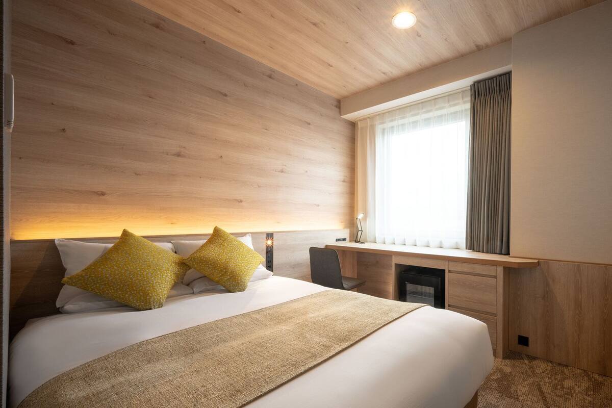 Une chambre d’hôtel avec une tête de lit en bois, un lit avec une literie blanche et deux oreillers jaunes, un bureau avec une lampe et une fenêtre avec des rideaux.
