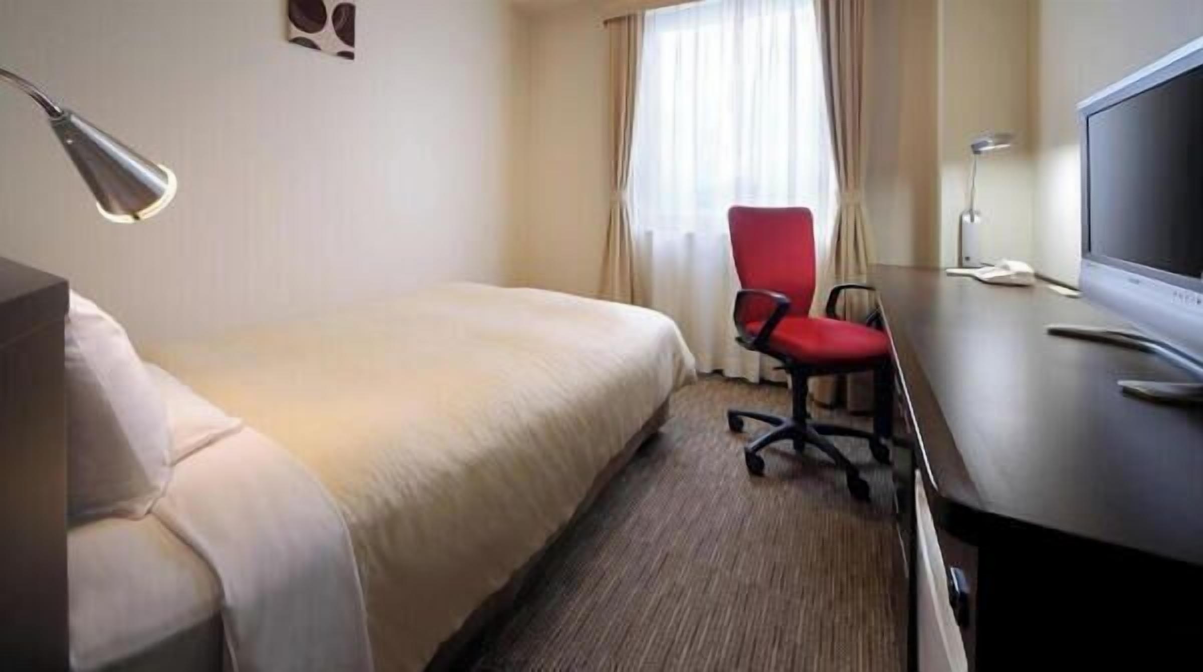 A hotel room with a bed, a desk with a chair, a television, and a window with curtains.