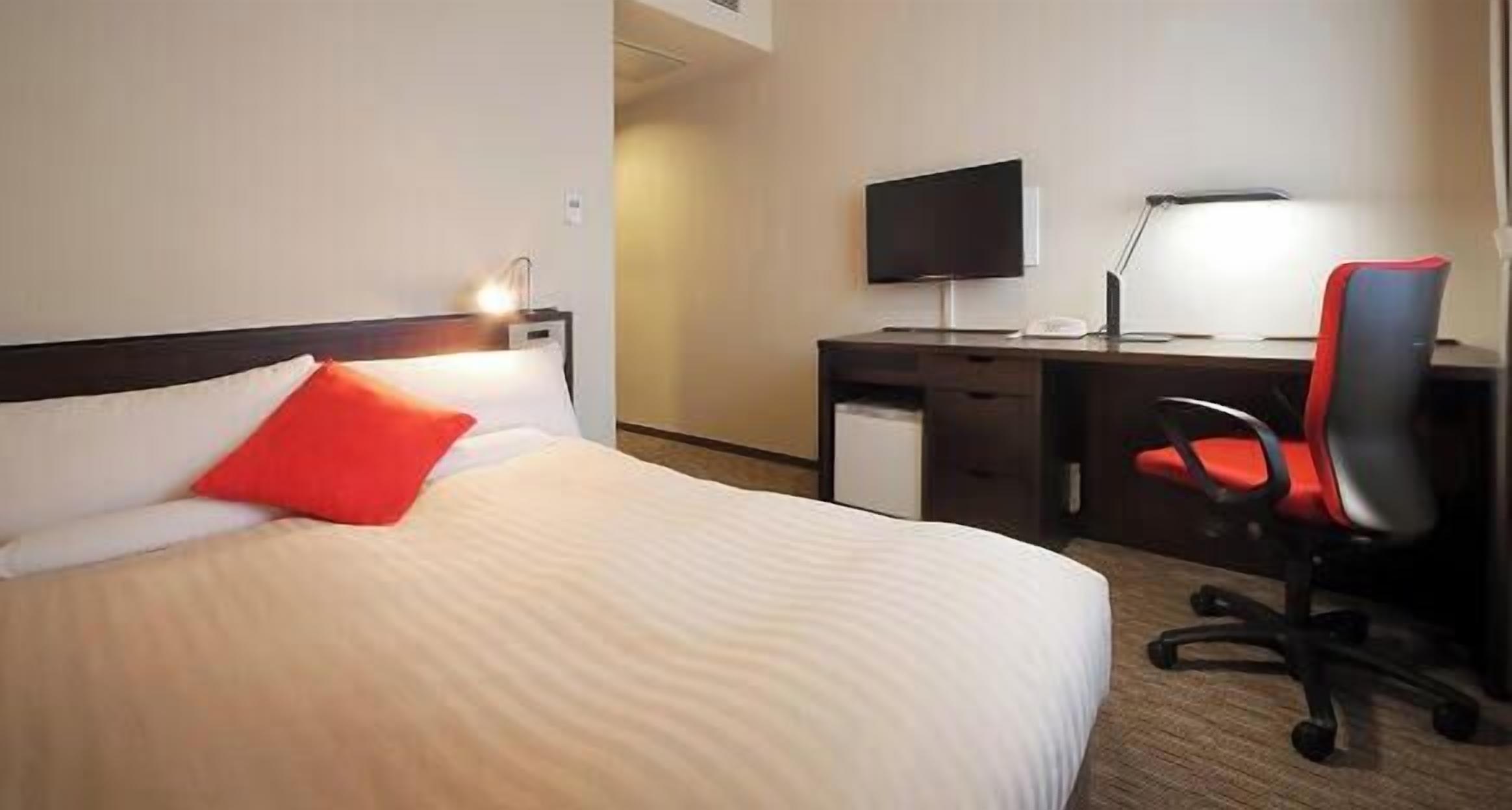 A hotel room with a bed, a desk, a chair, a TV, and a lamp.