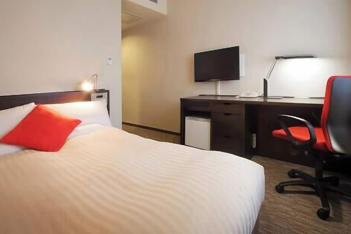A hotel room with a bed, a desk, a chair, a TV, and a lamp.