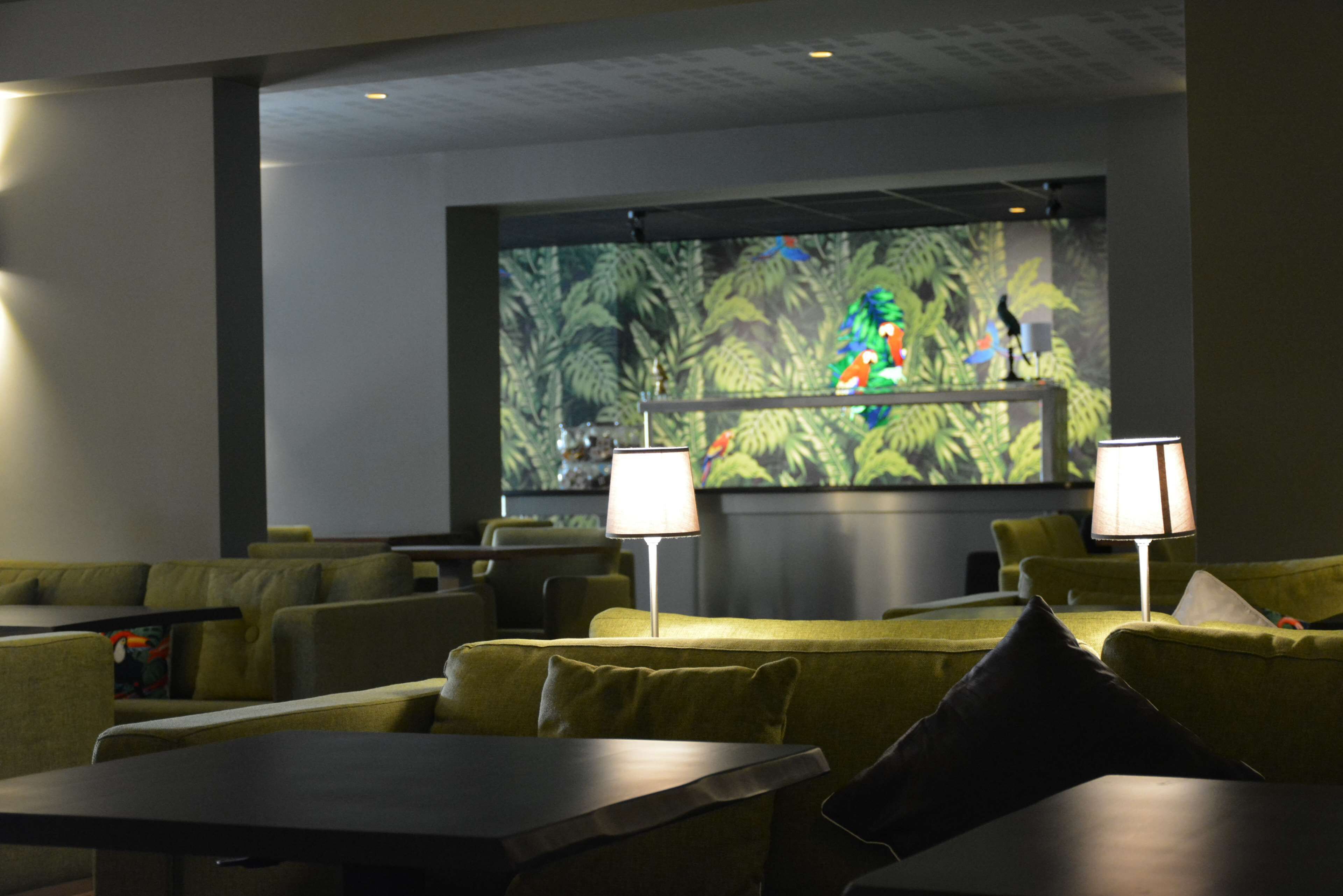 Un hall de hotel con sofás verdes, mesas y dos lámparas. Una pantalla grande muestra una imagen colorida.