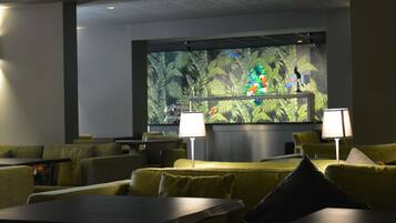 Un hall de hotel con sofás verdes, mesas y dos lámparas. Una pantalla grande muestra una imagen colorida.