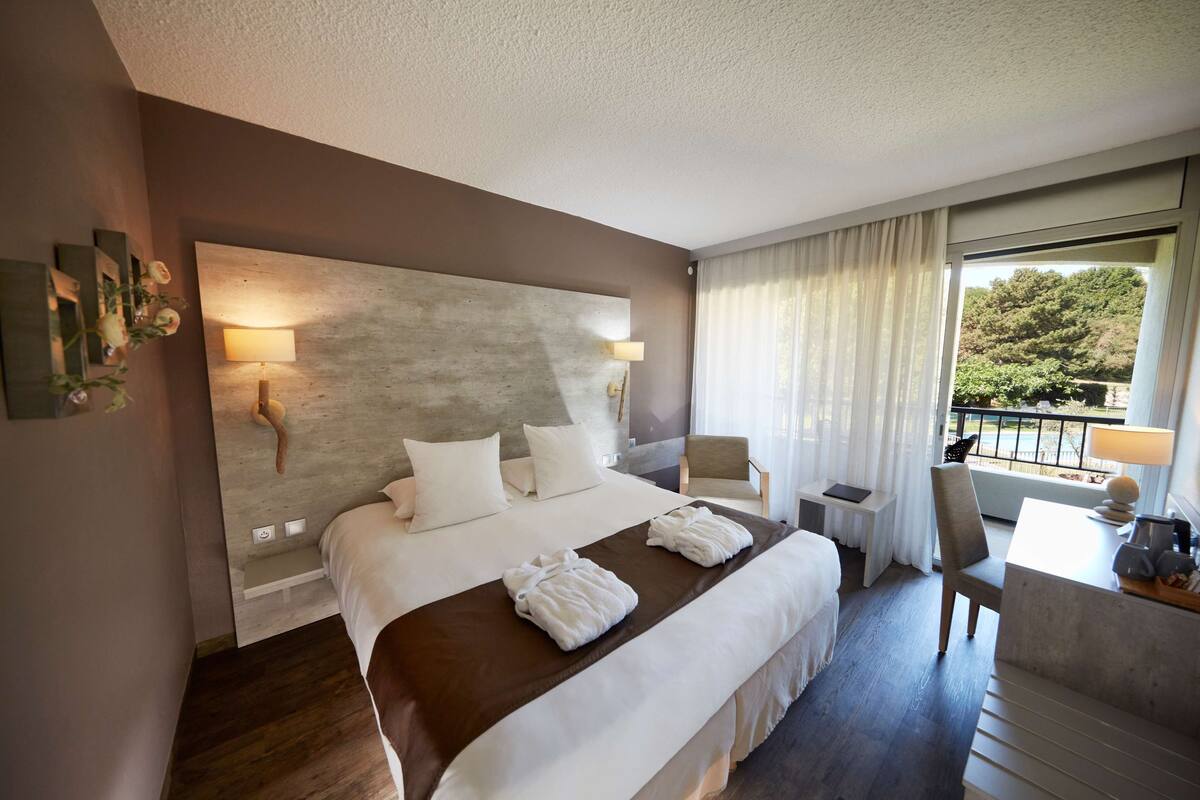 Habitación de hotel con una cama grande, un escritorio con silla, una mesita y vistas al exterior con zonas verdes.