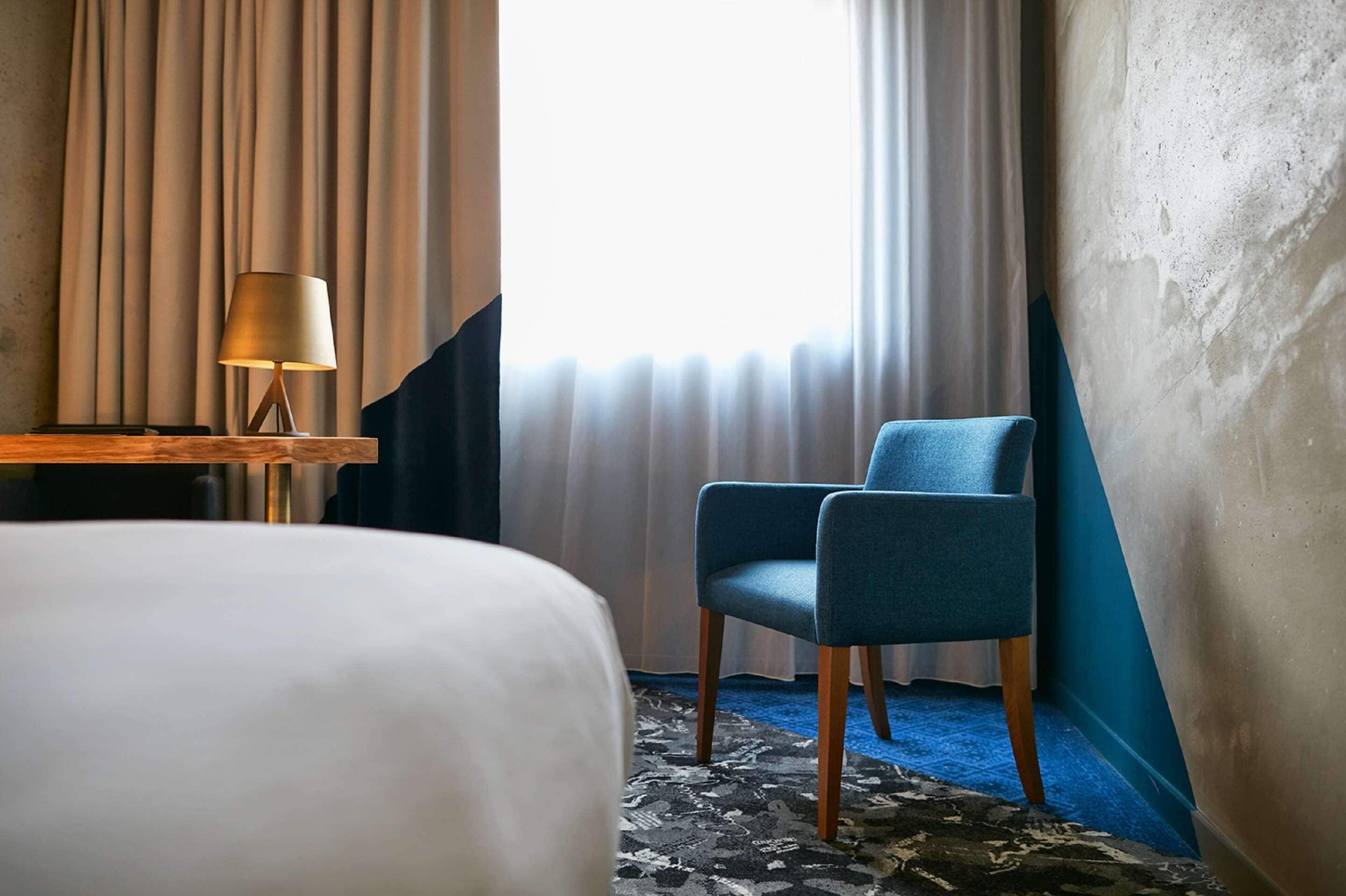 A hotel room with a bed, a chair, a lamp, and a window with curtains.