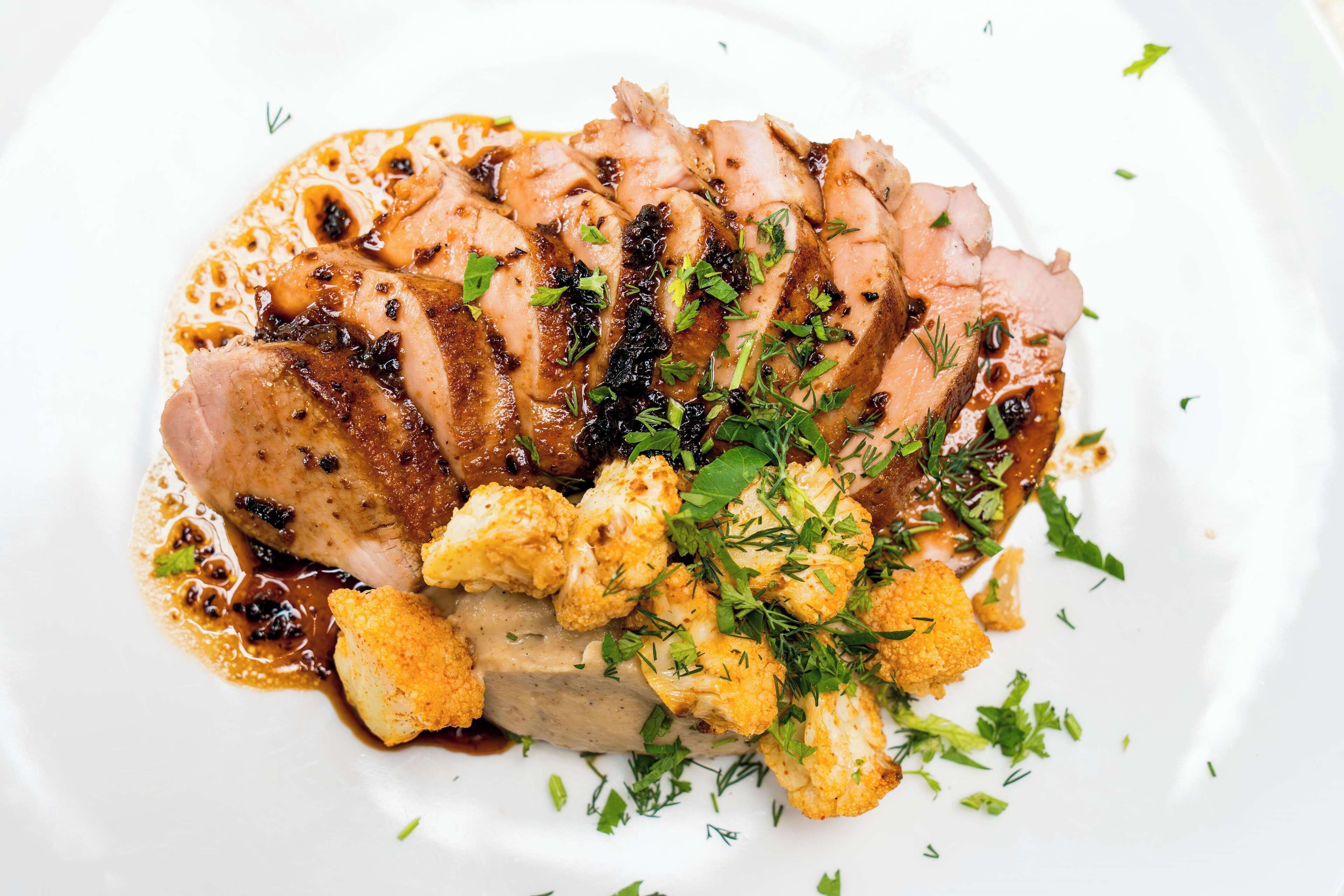 Ein Teller mit geschnittenem Fleisch, das eine dunkle Glasur hat, garniert mit Kräutern und dazu ein Beilagensalat aus Blumenkohl.