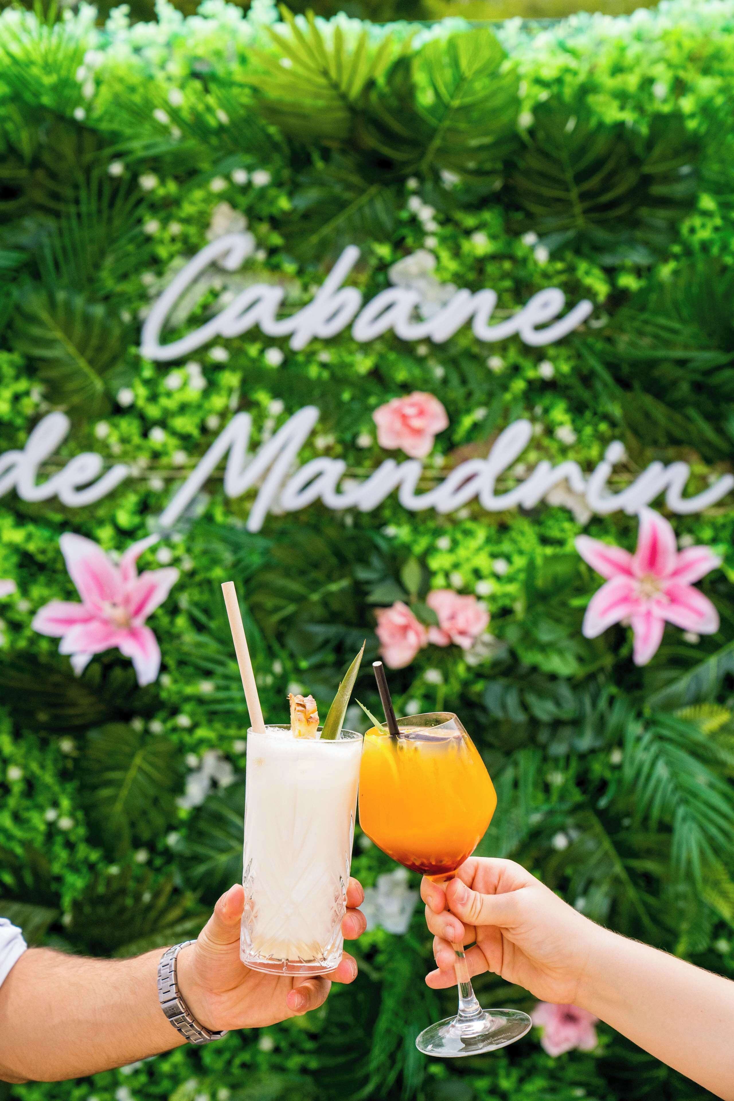 Two people holding drinks in front of a lush green wall with a sign that reads "Capone de Maindrin".