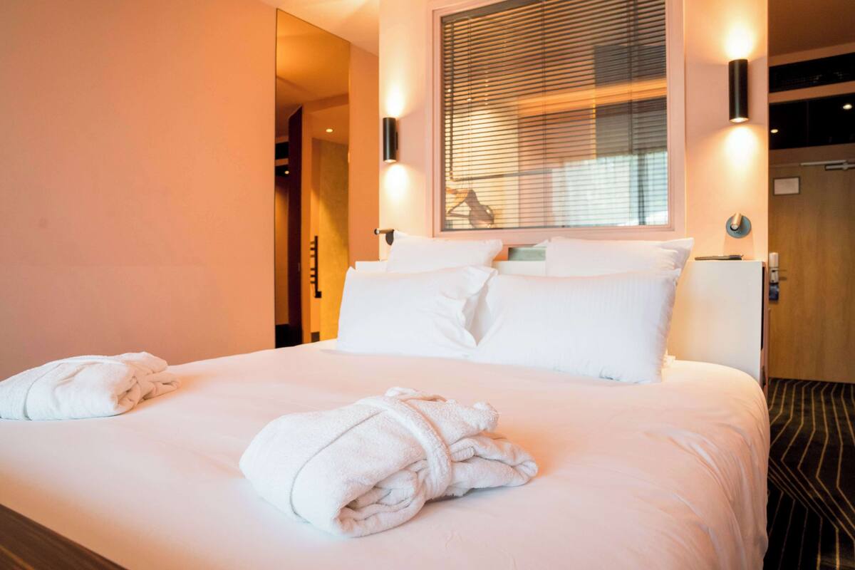 Una habitación de hotel con una cama bien hecha, dos lámparas fijadas a la pared y un ventanal con persianas.