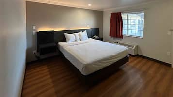 A bedroom with a bed, a bedside table, a nightstand, a window with curtains, and a wall-mounted heater.