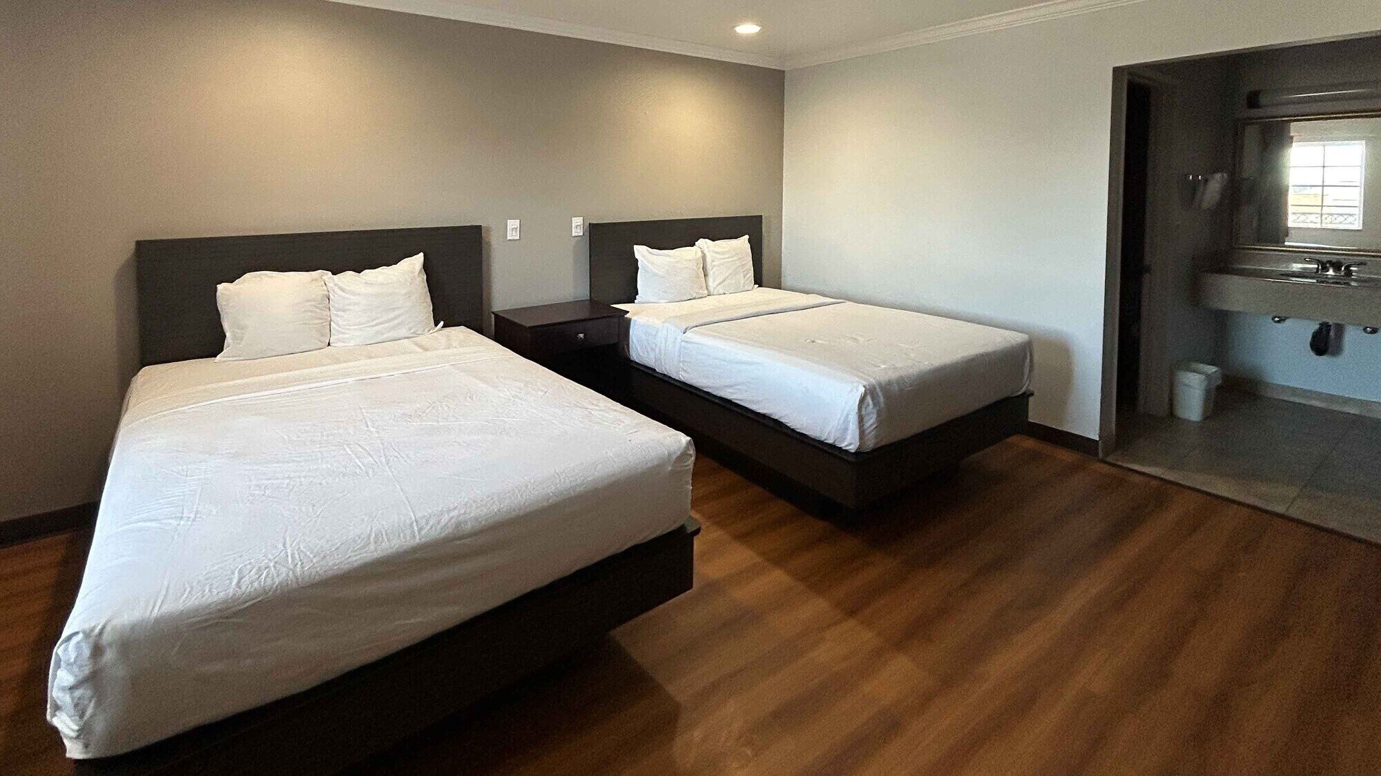 Habitación de hotel con dos camas, suelo de madera, baño pequeño con espejo y ventana.