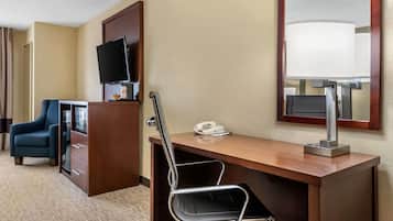 A hotel room with a wooden desk, a black office chair, a television mounted on the wall, a telephone, and a lamp.