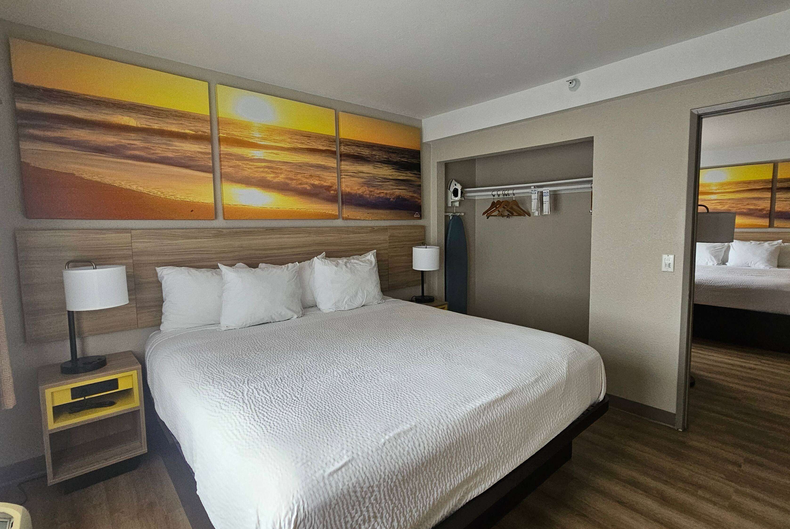Habitación de hotel con una cama grande, dos mesitas de noche, un armario y una pared con tres cuadros de atardecer.
