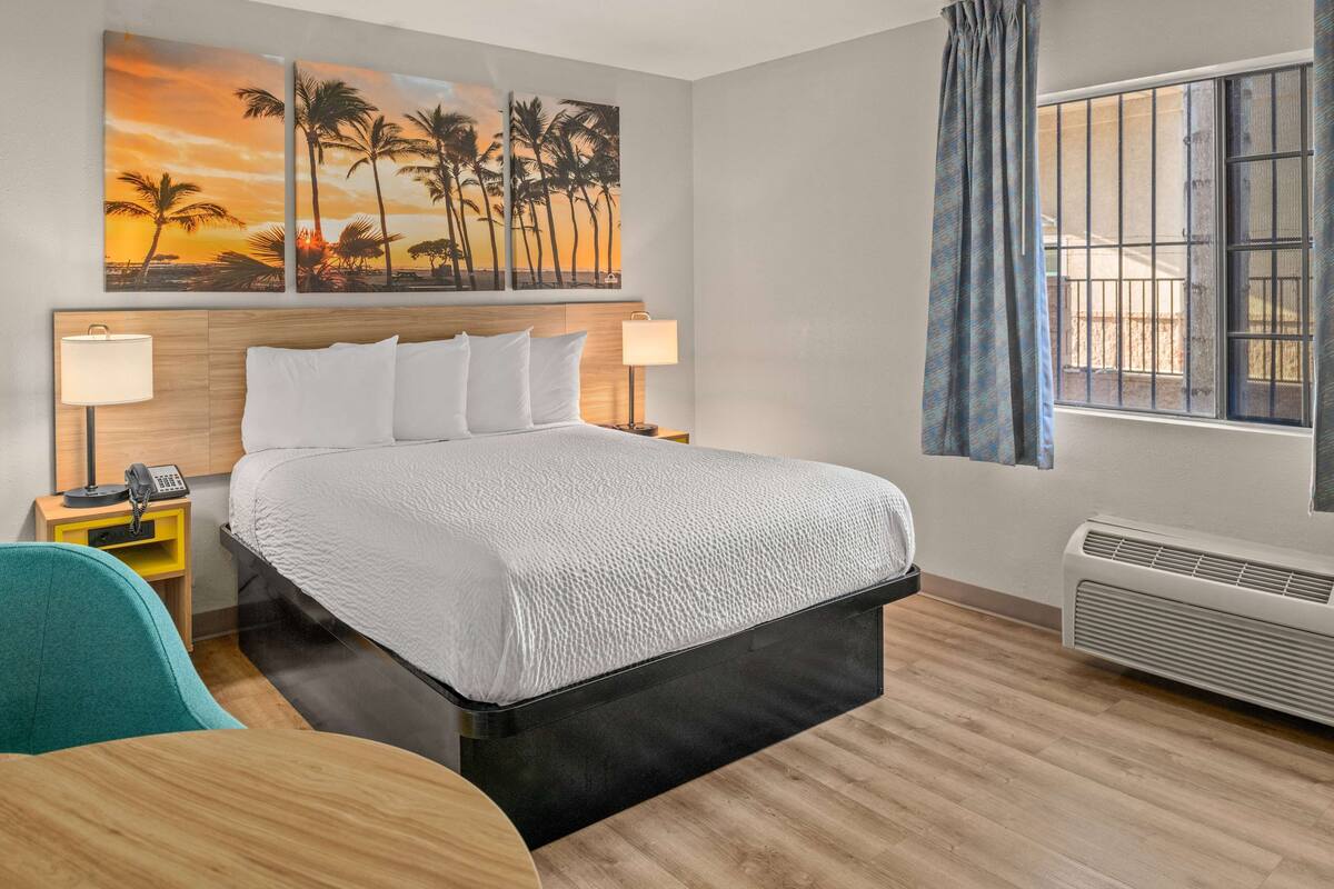 Une chambre d’hôtel comprenant un lit, deux tables de chevet avec des lampes, un bureau, une chaise, une grande fenêtre avec vue et un mur orné d’un tableau représentant un coucher de soleil.