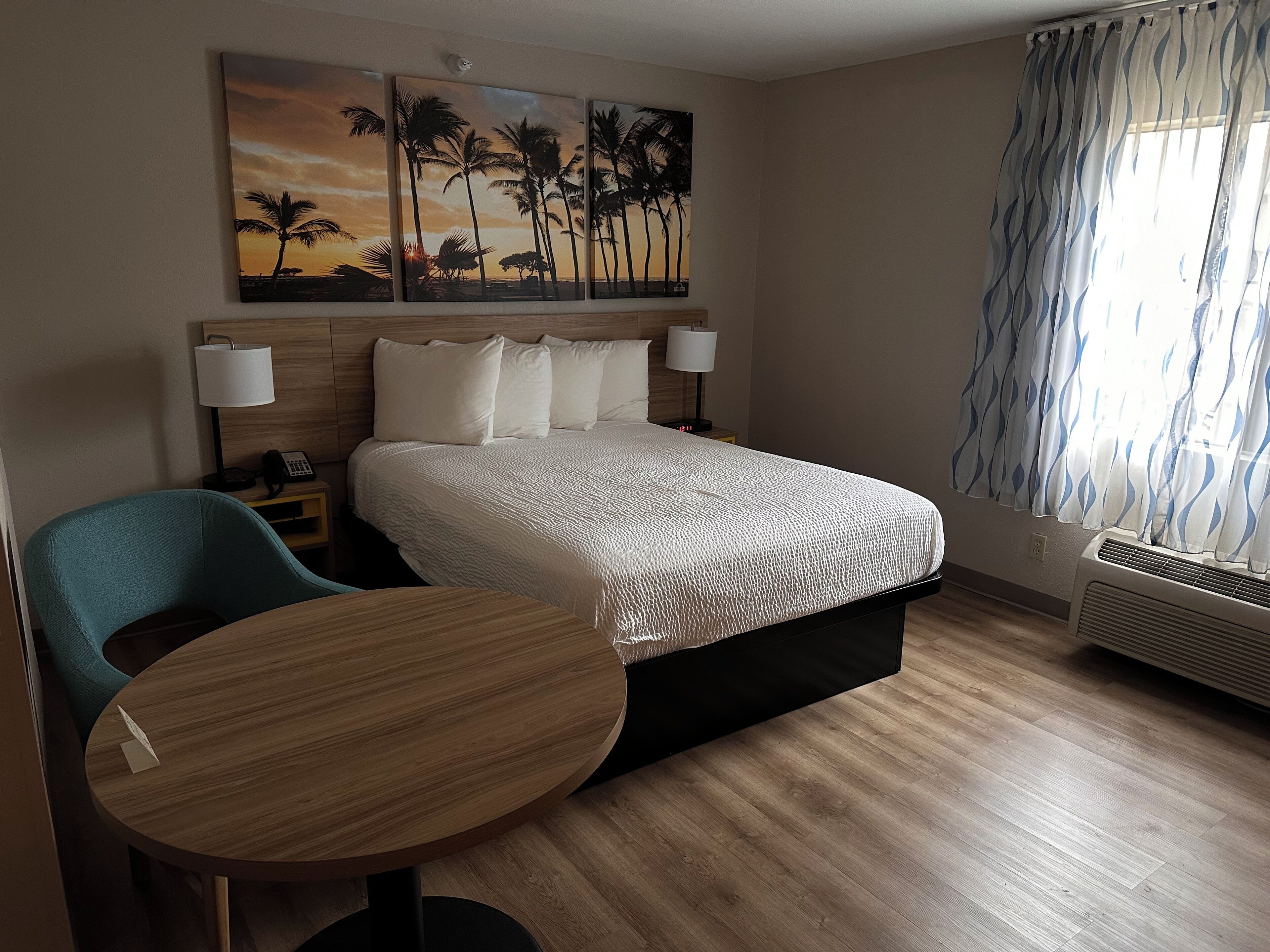 Habitación de hotel con cama, lámparas de noche, escritorio, silla, un cuadro de palmeras y una ventana con cortinas.
