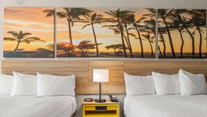 A hotel room with two beds, a wooden headboard, a nightstand with a lamp, and a wall-mounted painting of palm trees at sunset.